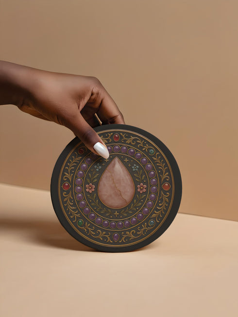 Round ceramic drink coaster featuring a detailed rose quartz-inspired teardrop surrounded by floral and gemstone baroque patterns. Black background with jewel-toned accents and gold scrollwork. The coaster has a non-slip cork base and measures approximately 4 inches in diameter. Perfect for hot or cold beverages, this witchy decor piece enhances any coffee cart, altar, or side table. Suitable for use in whimsigoth, gothic, or celestial-themed spaces.